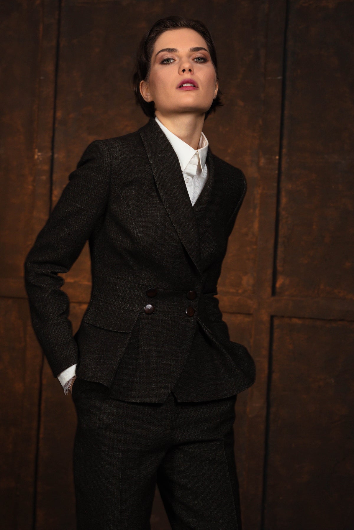 A woman wearing a tailored dark charcoal double-breasted blazer over a white collared shirt, paired with matching dark trousers, posed against a dark brown wood-paneled background.