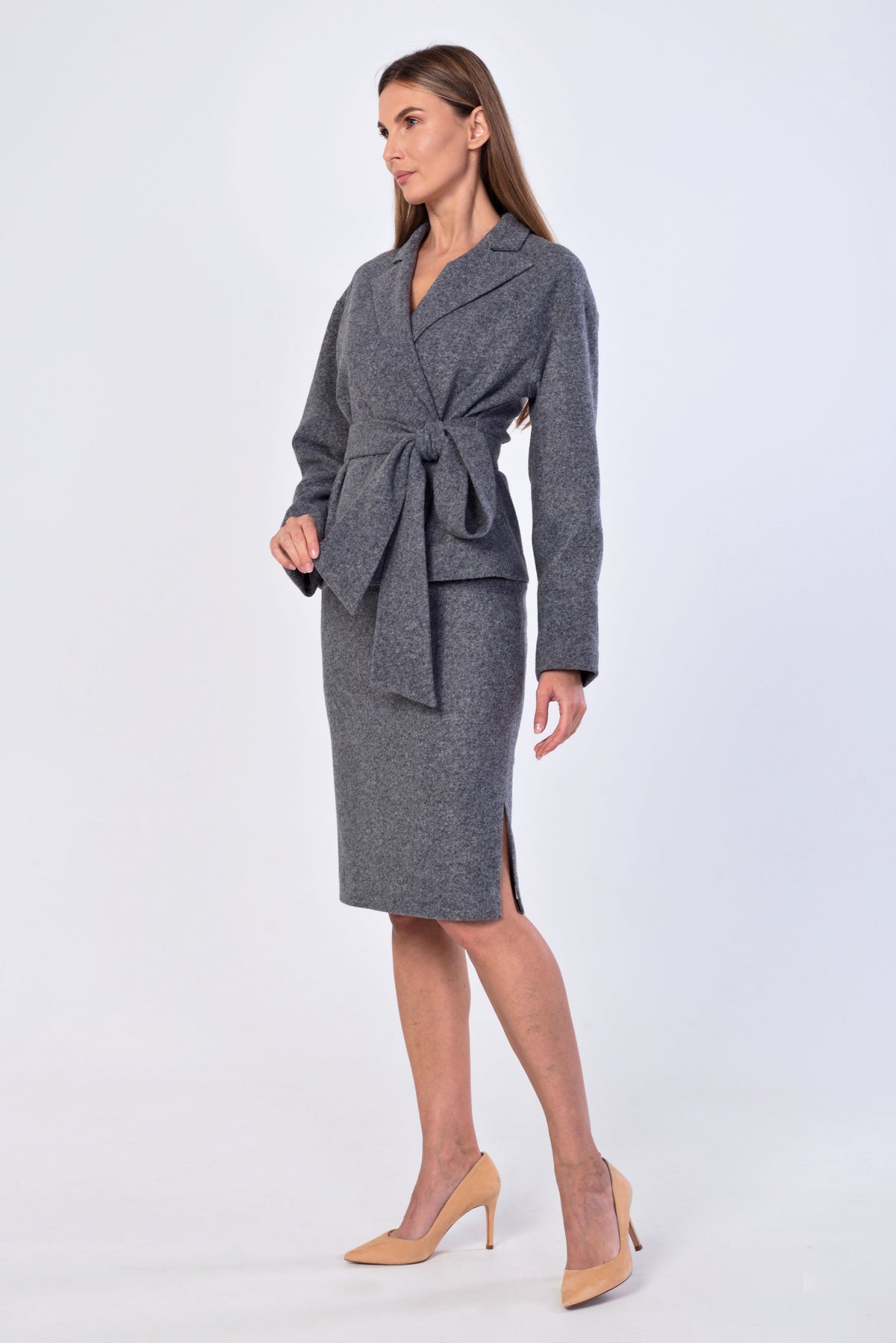 A woman modeling a gray textured wrap jacket with a tie waist, with a matching knee-length skirt, paired with beige high heels, against a light gray studio background.