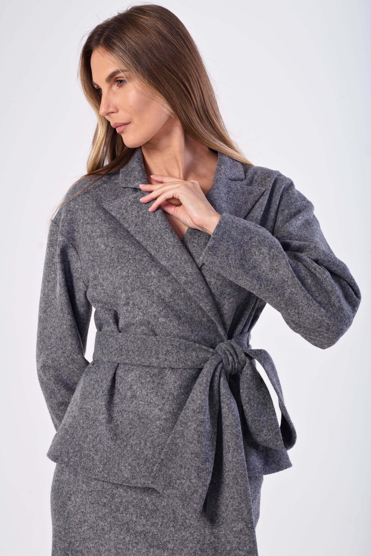A woman wearing a gray wool wrap jacketwith a matching tie belt, photographed against a plain light gray studio background.
