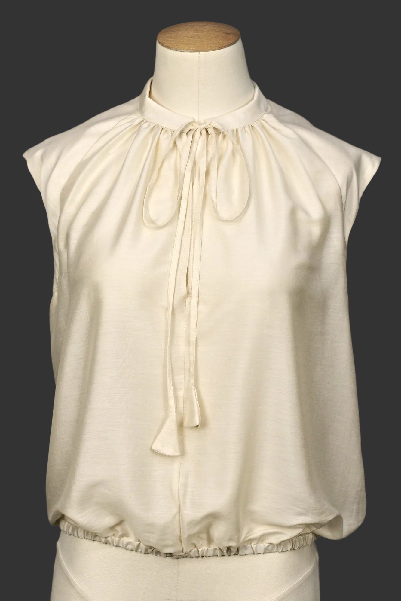 Close‑up of ivory sleeveless silk–viscose blouse showing front tie detail and soft sheen, on a mannequin.