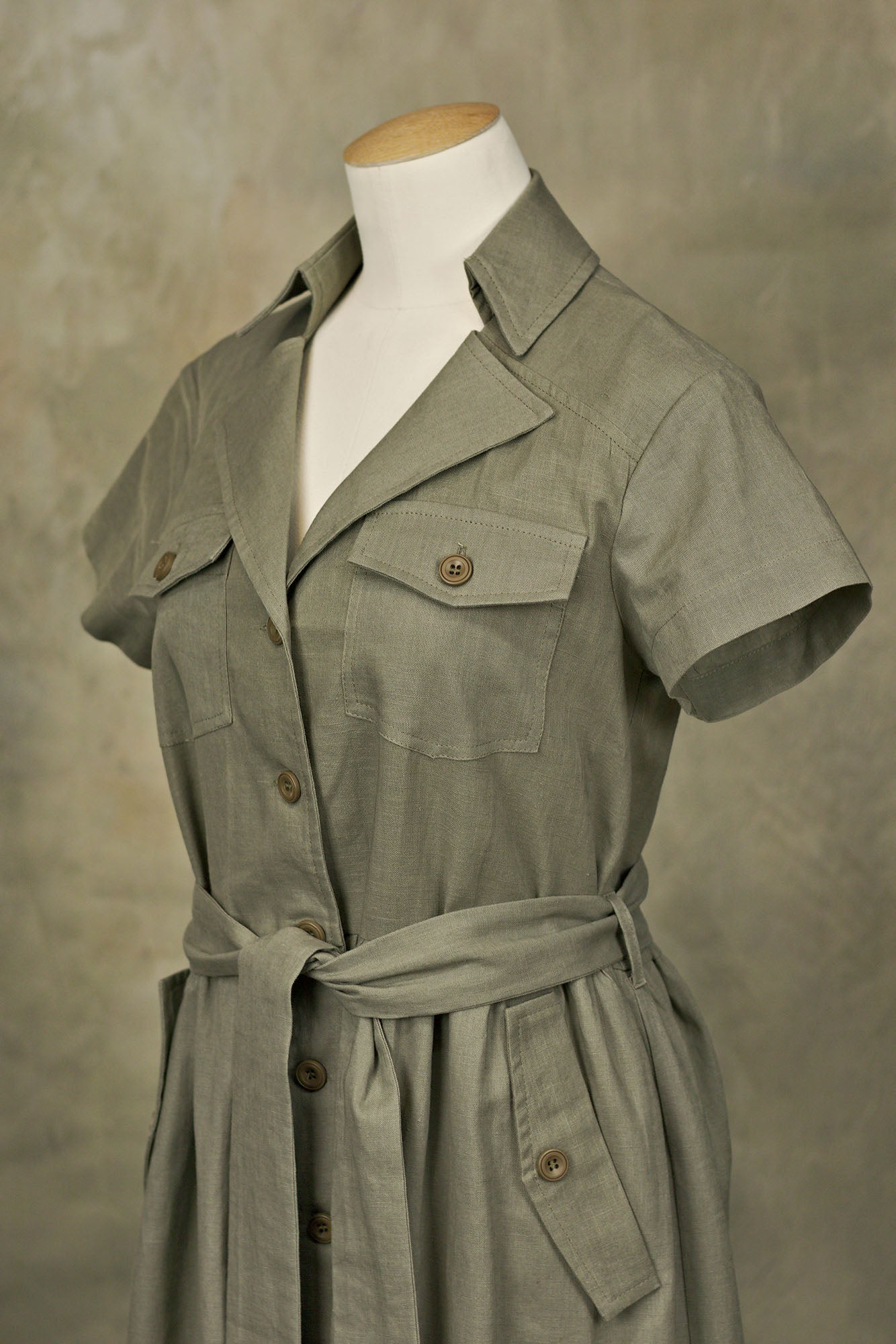Olive green linen button‑down shirt-dress with collared neckline, chest pockets, and belted waist, on a mannequin, angled front studio shot.