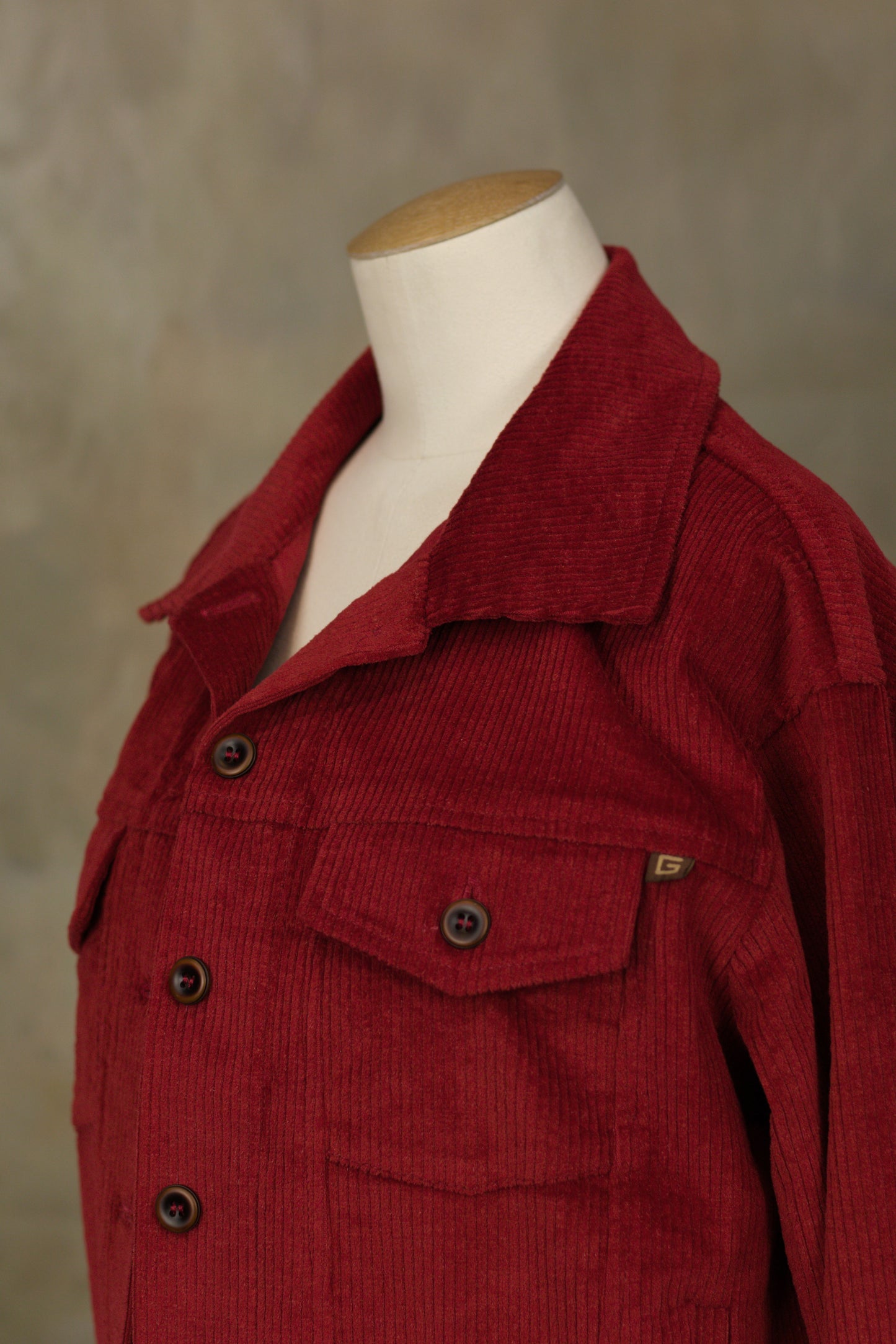 Close-up detail of a dark red corduroy jacket showing the vertical ribbed texture, chest flap pocket, and branded logo tag.