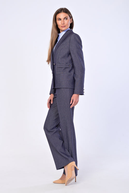 A woman modeling a tailored two-piece suit in a slate-blue, featuring a double-breasted blazer and matching straight-leg trousers, styled with a light blue collared shirt and beige high-heeled pumps against a light gray studio background.