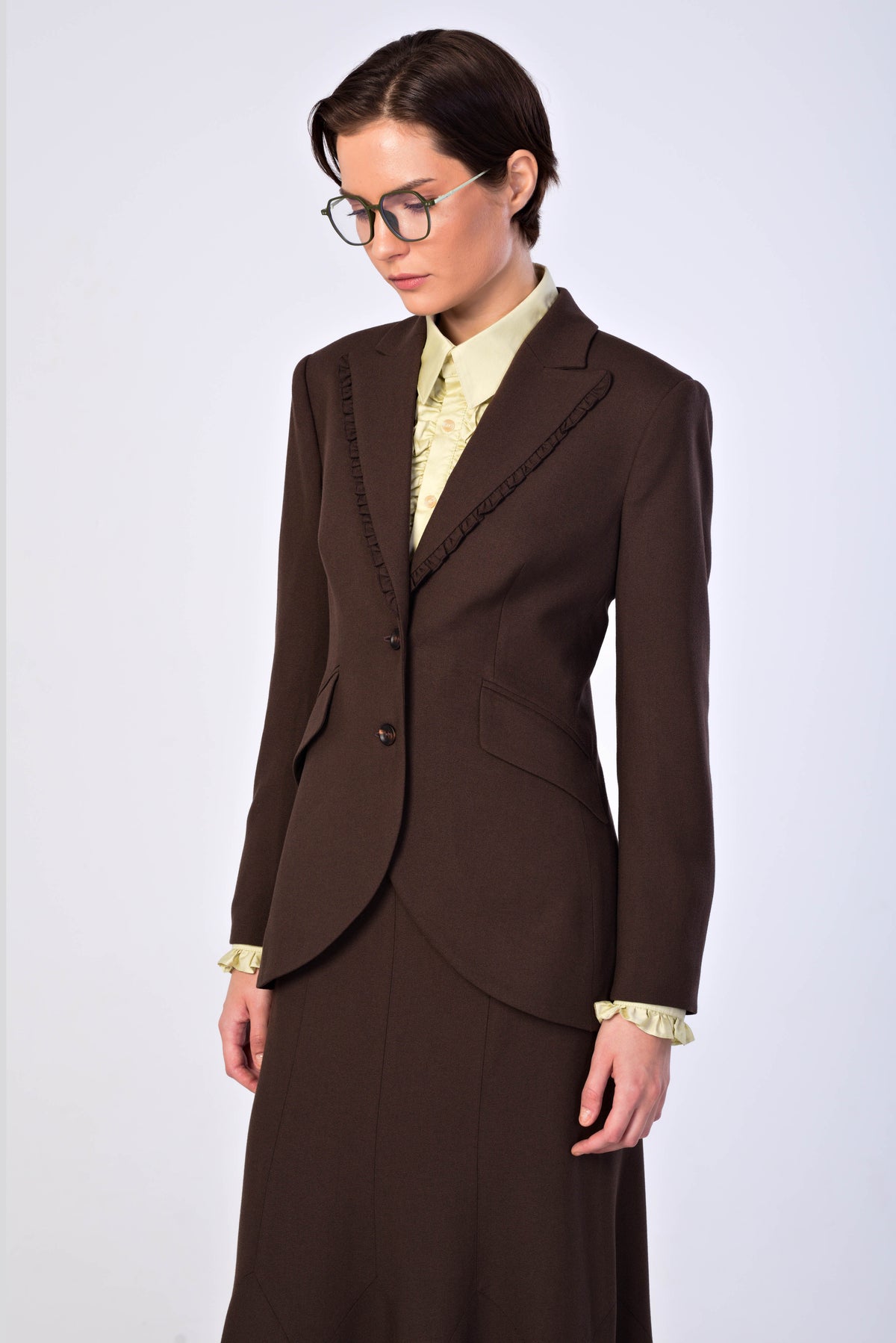 A woman wearing a dark brown tailored suit with a matching skirt, a pale yellow patterned blouse, and glasses, photographed against a light gray studio background.