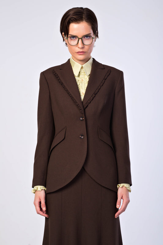 A model wearing a dark brown tailored blazer with decorative ruched detailing along the lapels, paired with a light yellow patterned shirt and a matching dark brown skirt, set against a plain light gray background.