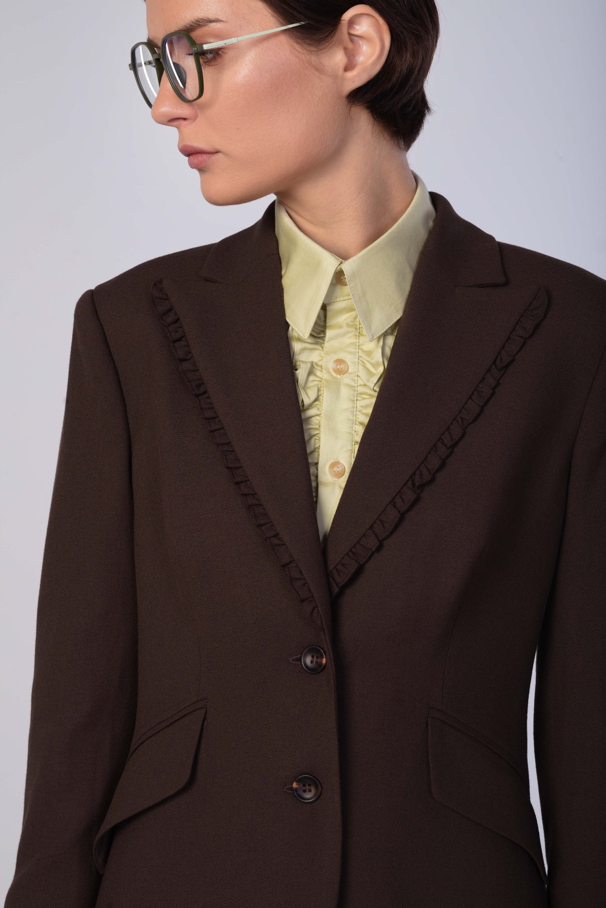 A close-up of a model wearing a dark brown tailored blazer with decorative ruffle trim along the lapels, paired with a pale yellow ruffled shirt and glasses.