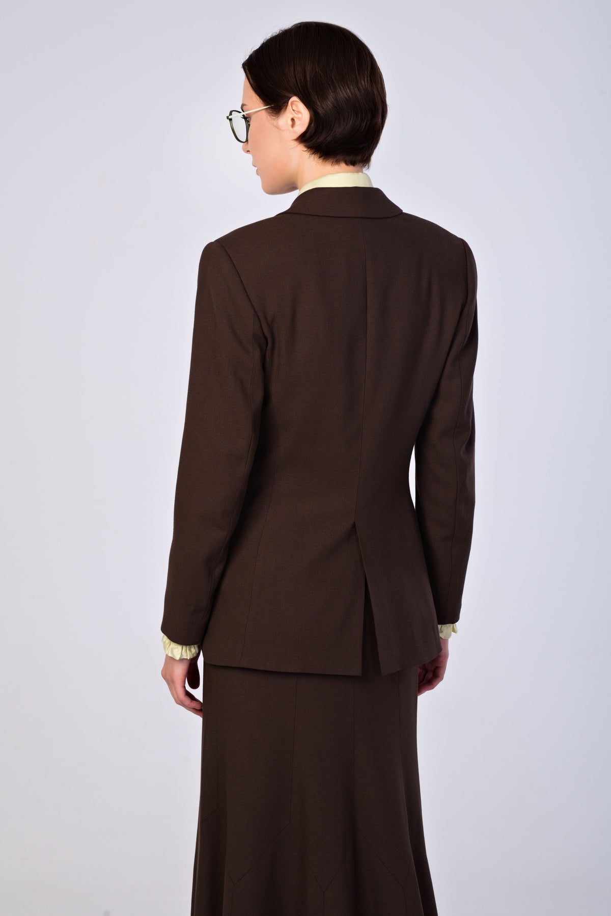 A model shown from the back wearing a dark brown tailored blazer and matching skirt, with a light-colored collared shirt visible at the neckline, set against a plain light gray studio background.