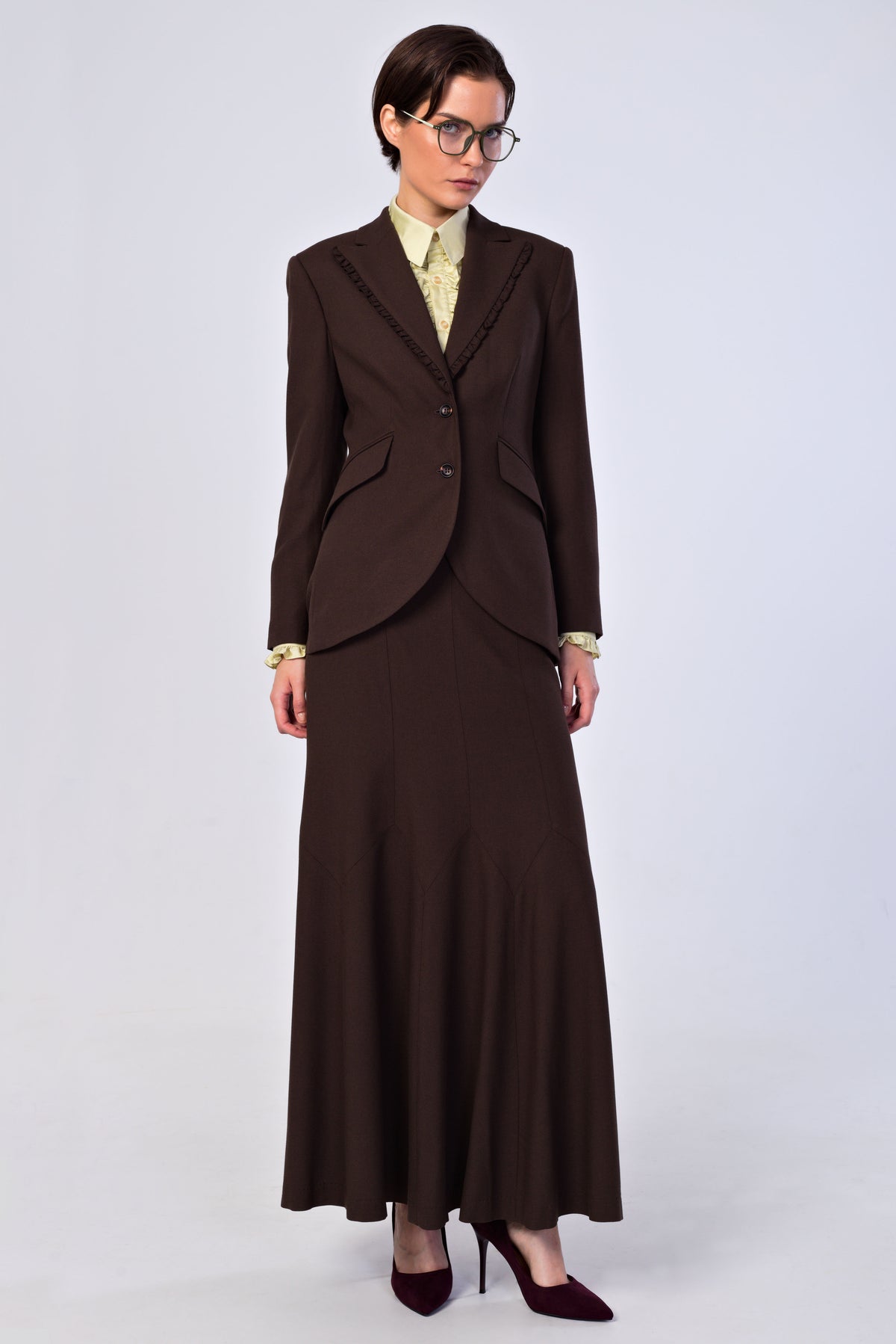 A model wearing a dark brown two-piece suit consisting of a single-breasted blazer and a matching long skirt, paired with a light yellow patterned shirt, glasses, and burgundy heels, against a light gray studio background.