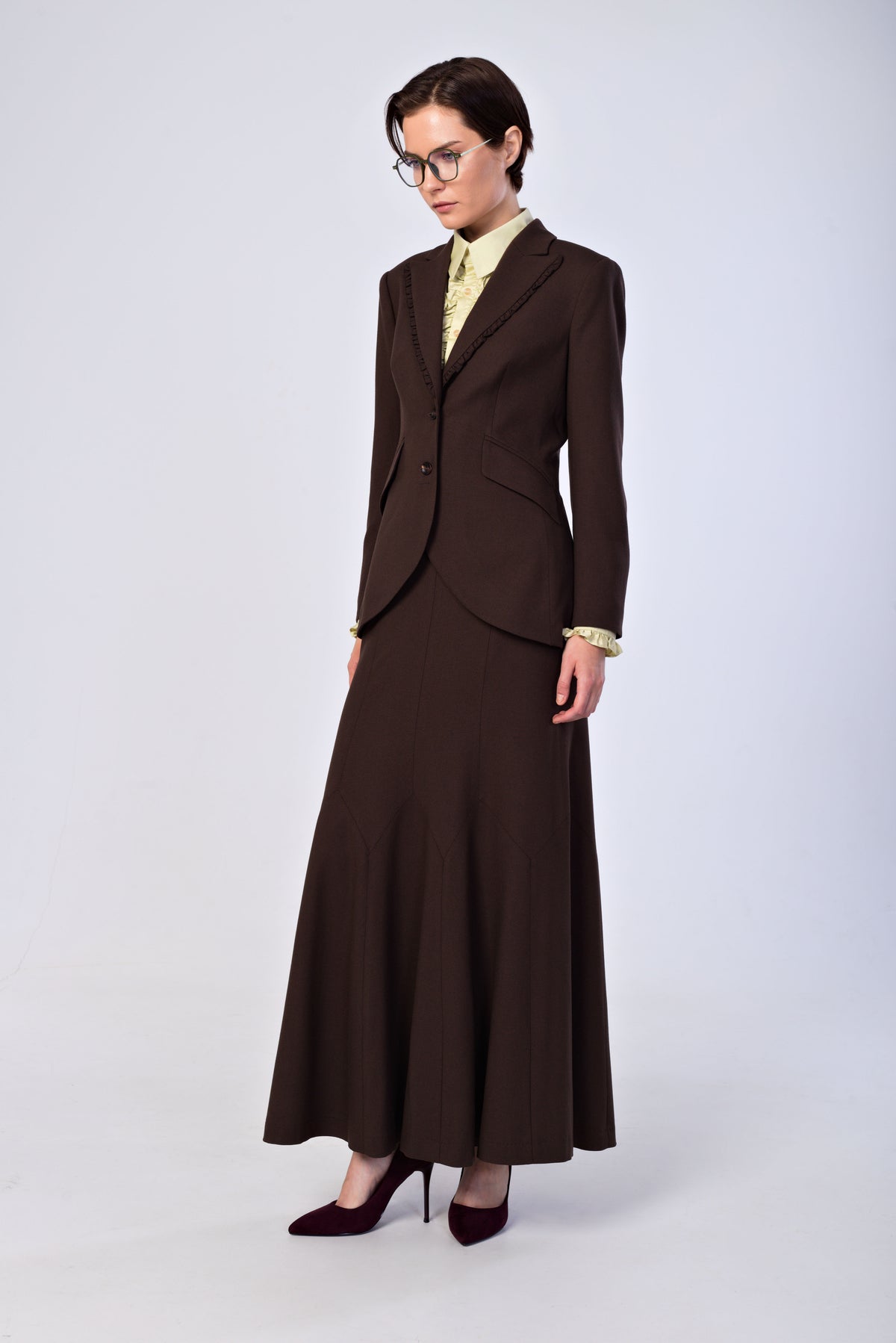 A model wearing a dark brown two-piece suit consisting of a tailored blazer and a long A-line skirt, styled with a light yellow patterned shirt, gold cufflinks, and dark burgundy heels against a light gray studio background.
