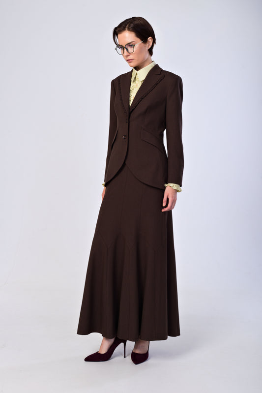 A model wearing a dark brown two-piece suit consisting of a tailored blazer and a long A-line skirt, styled with a light yellow patterned shirt, gold cufflinks, and dark burgundy heels against a light gray studio background.