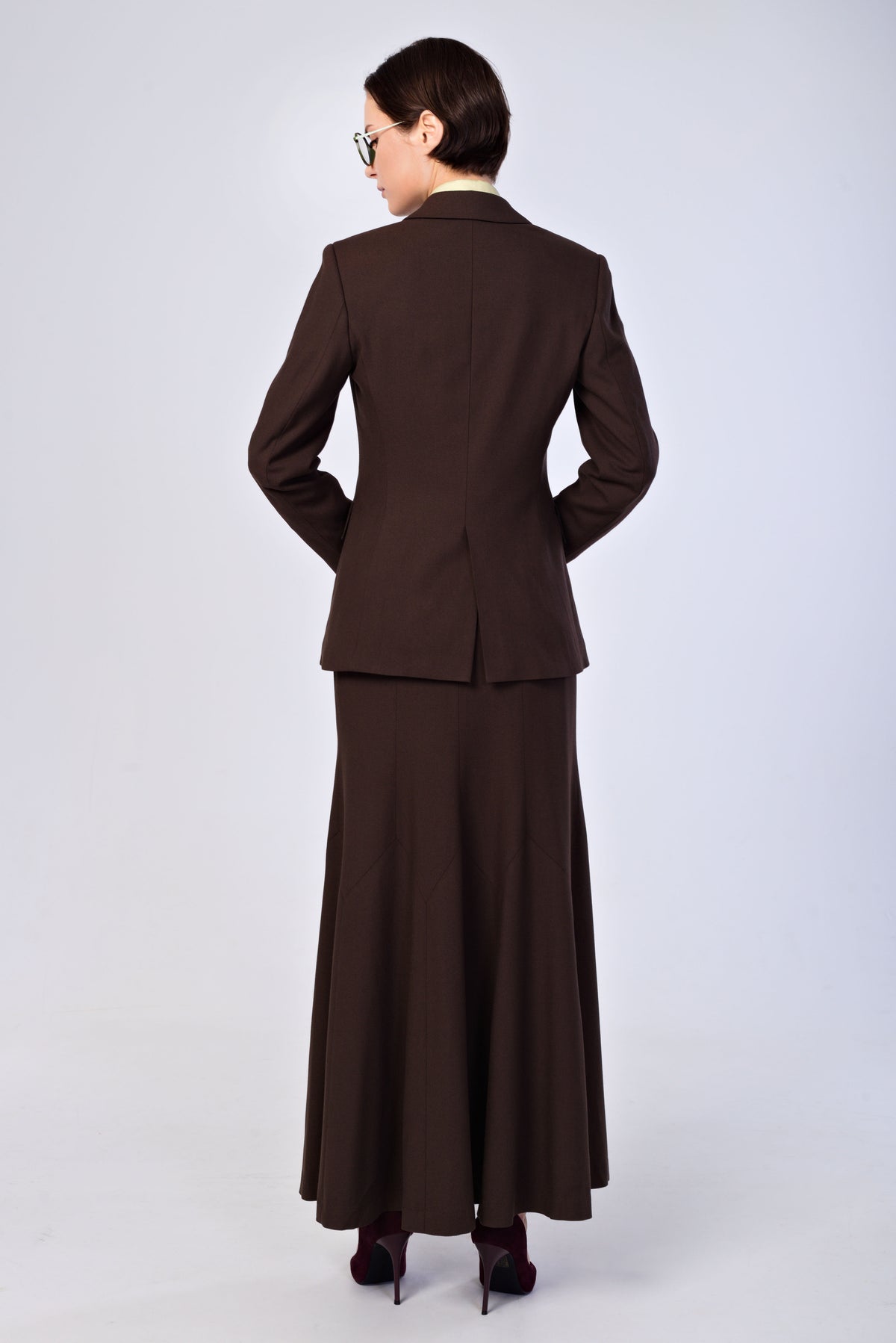 A woman shown from the back wearing a dark brown tailored suit with a long skirt and heels, photographed against a plain light gray studio background.