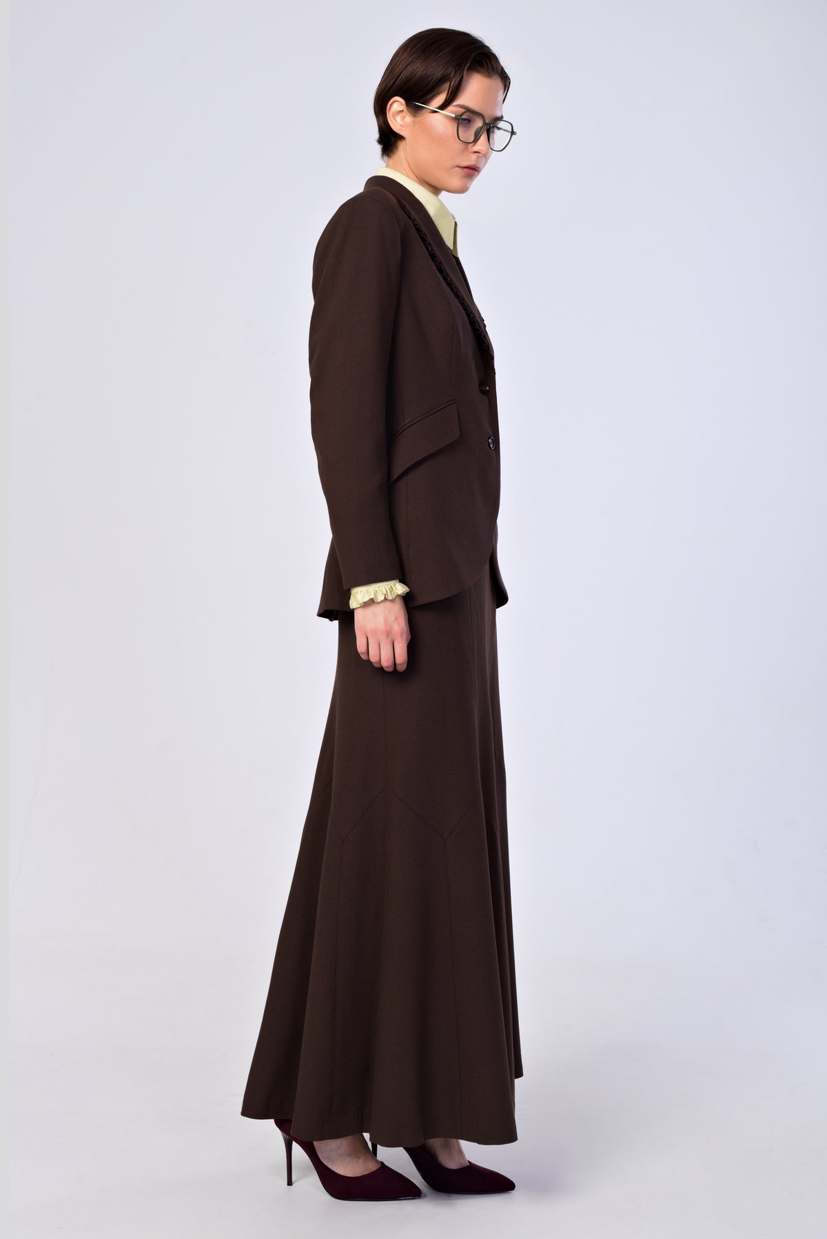 A model wearing a dark brown two-piece suit with a structured blazer and matching long skirt, styled with a cream collared shirt, glasses, and dark red heels against a light gray studio background.