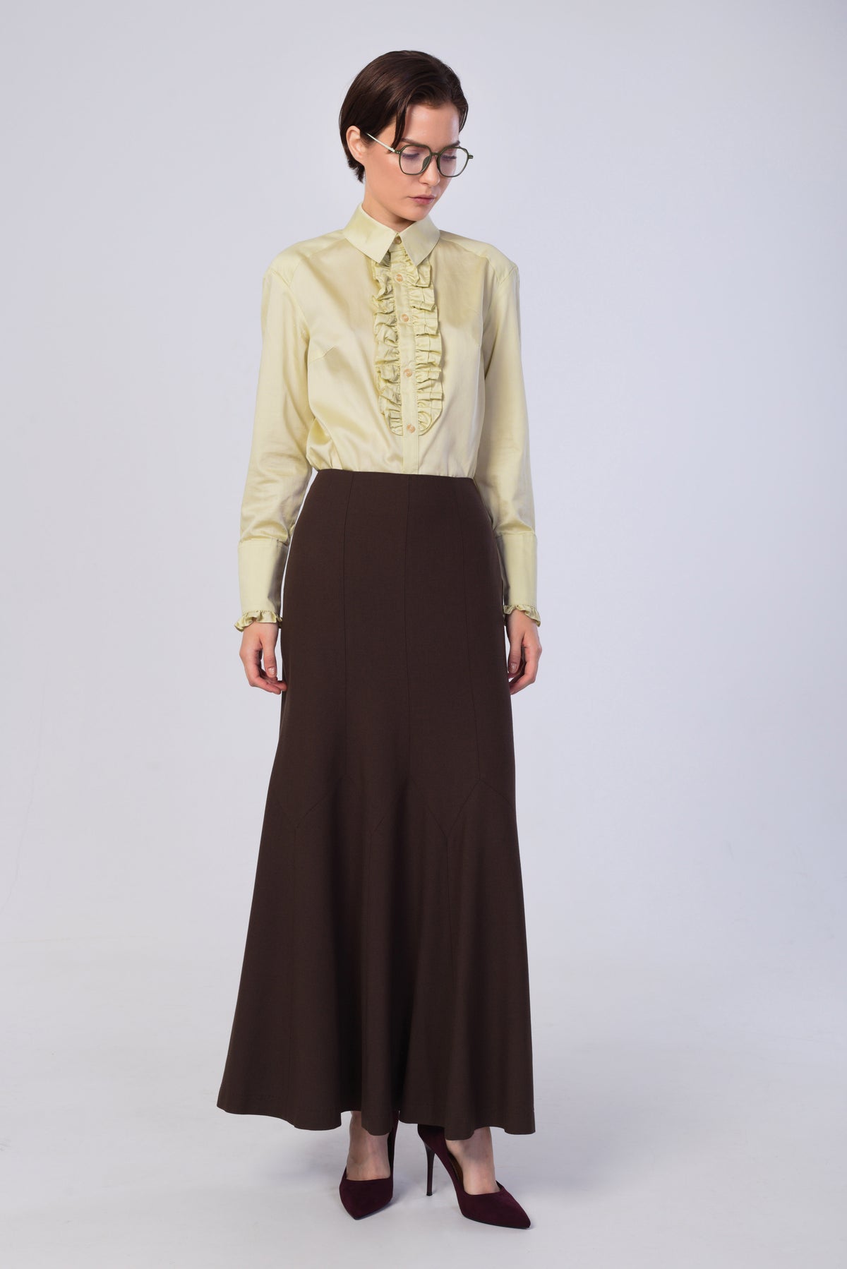 A model wearing a pale yellow long-sleeve blouse with a ruffled front and a dark brown high-waisted maxi skirt, paired with dark burgundy heels, against a light gray studio background.