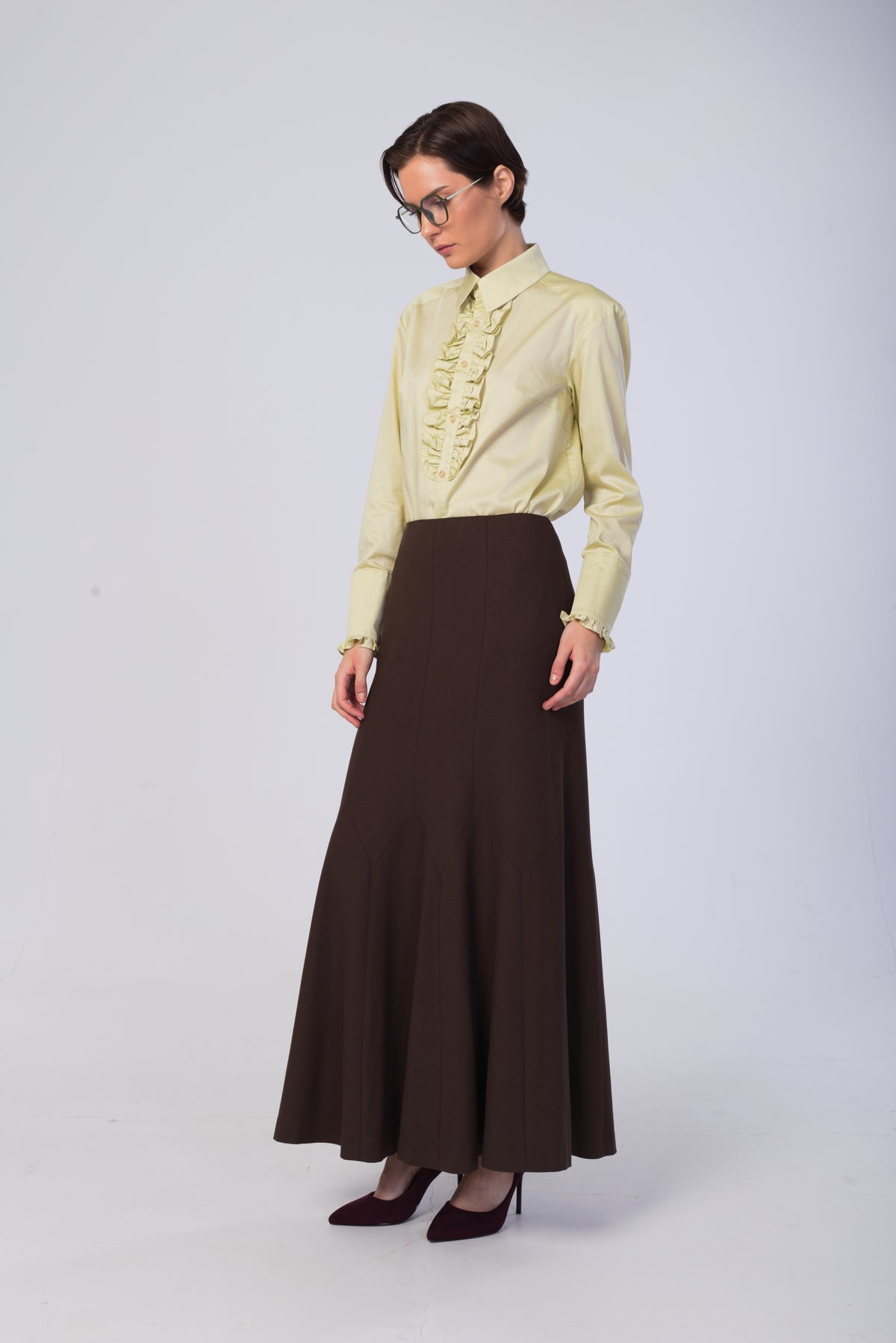 A woman modeling a light yellow long-sleeve blouse with a ruffled front and a dark brown high-waisted maxi skirt, paired with dark brown heels, against a light gray studio background.