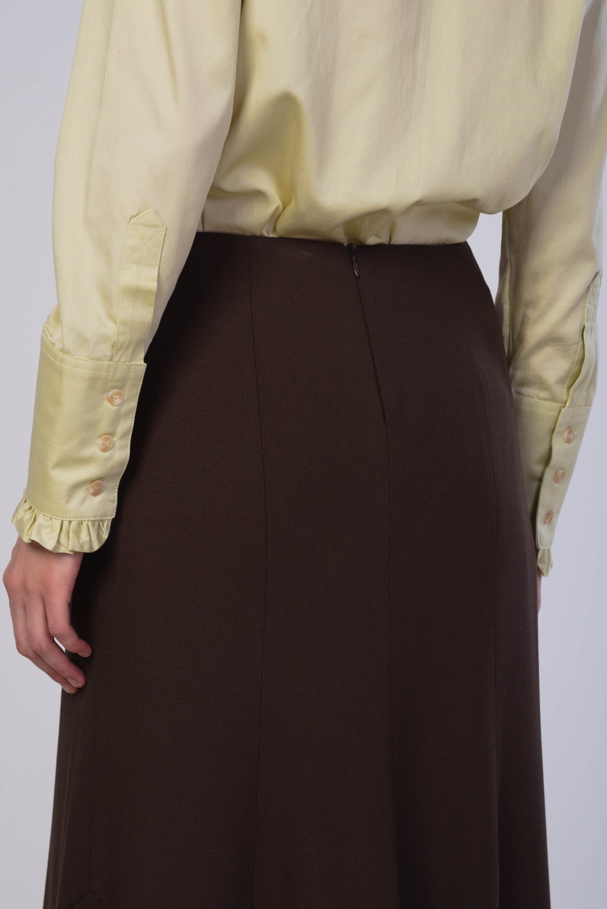 A close-up of a person wearing a dark brown skirt and a pale yellow satin blouse with ruffled cuffs, shown from the back against a light gray studio background.