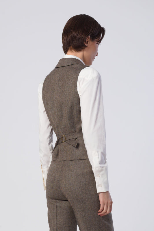 A model shown from the back wearing a tailored brown tweed vest and matching trousers over a white long-sleeve shirt, against a light gray studio background.