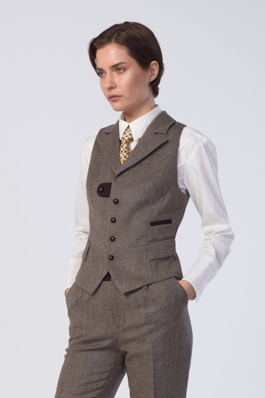 A woman modeling a tailored two-piece suit set consisting of a brown tweed vest and matching trousers, worn over a white collared shirt with a patterned tie, against a light gray studio background.