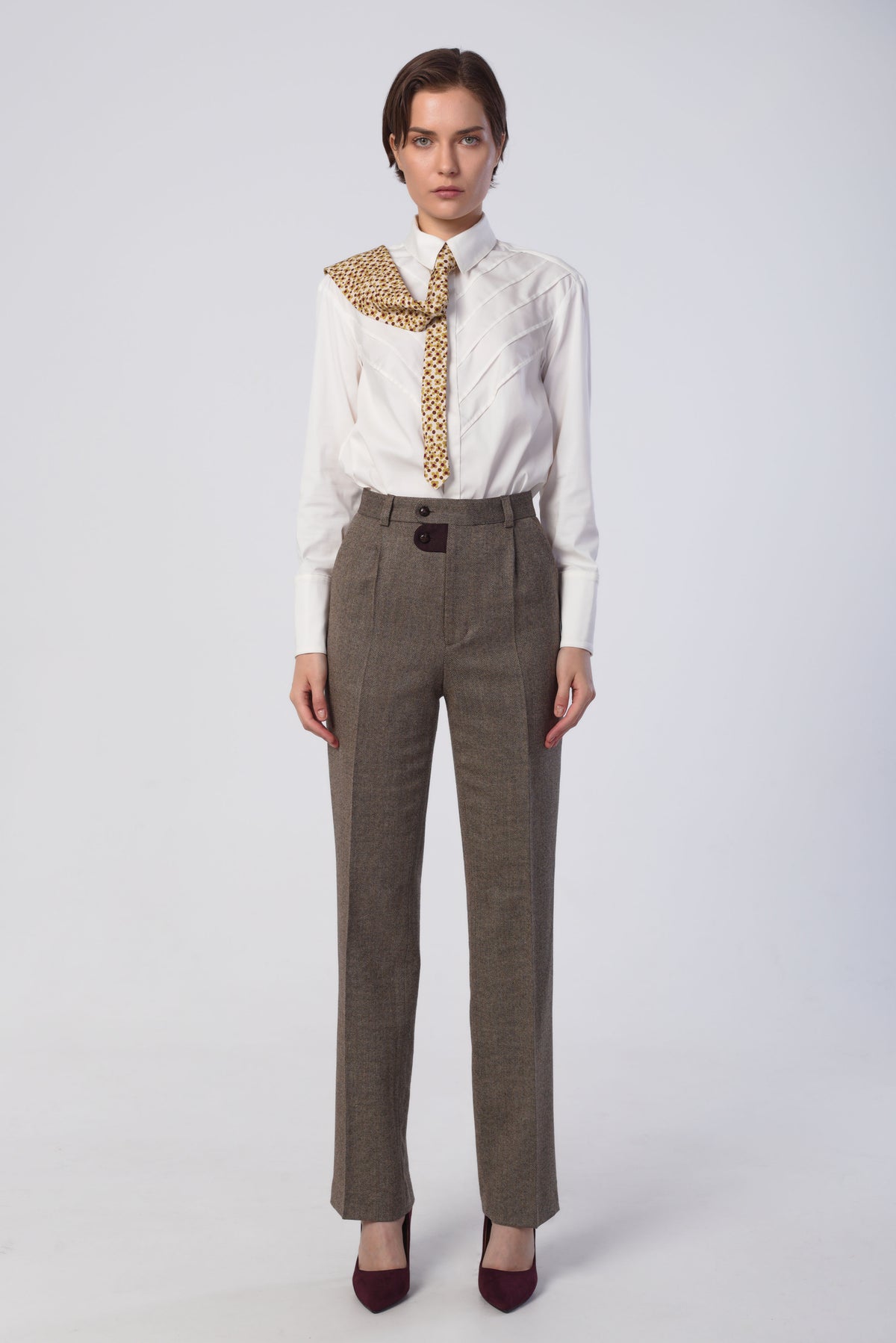 A model stands against a light gray studio background wearing a white long-sleeve shirt, a matching gold patterned necktie, high-waisted brown trousers, and dark burgundy heels.