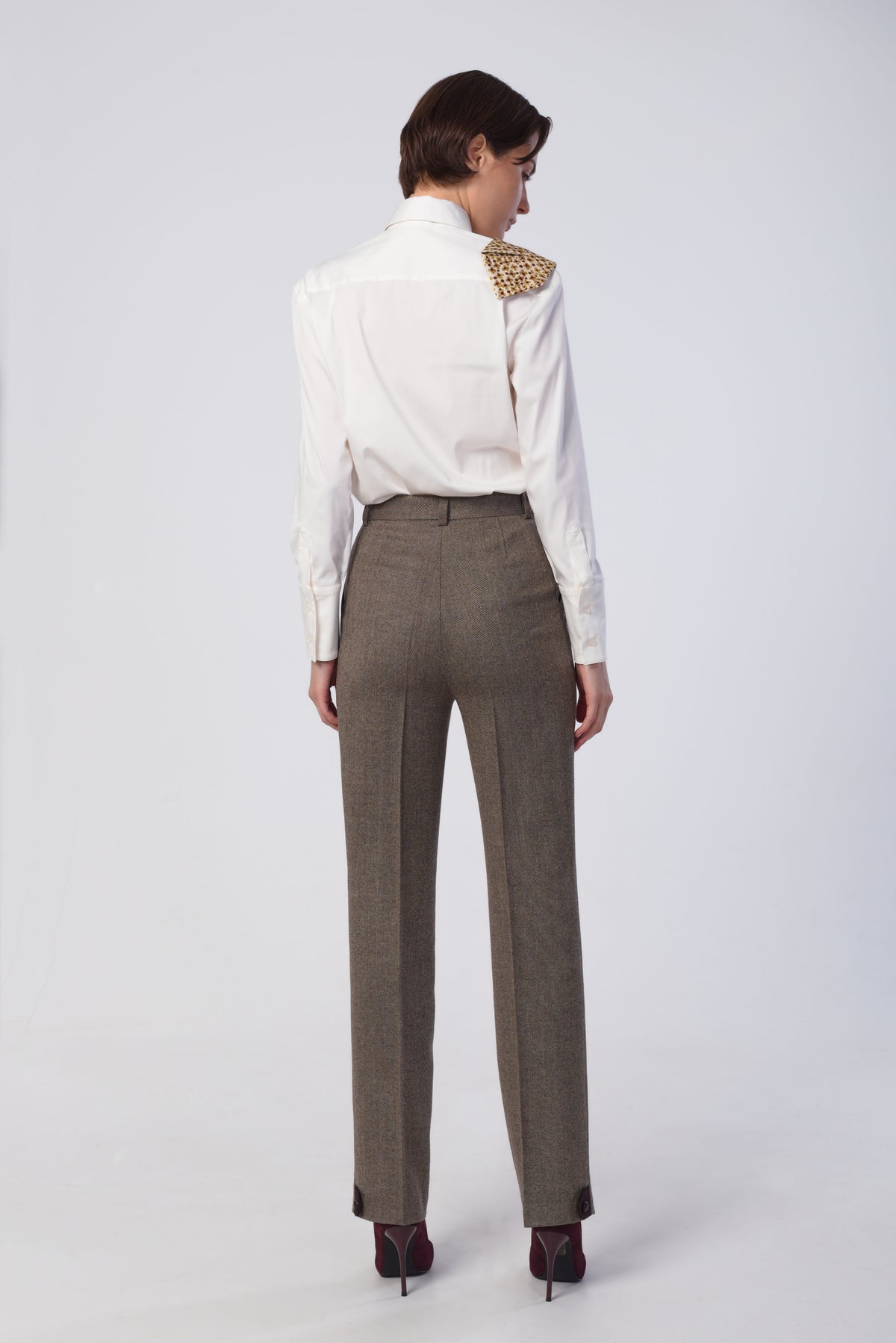 A model shown from the back wearing a white long-sleeve shirt paired with high-waisted brown trousers and dark red heels, against a light gray studio background.
