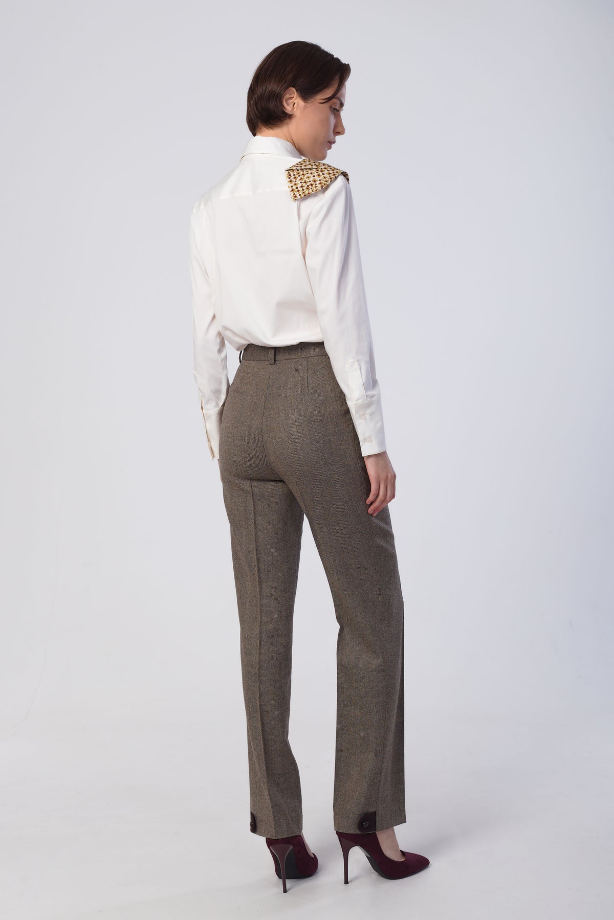 A woman shown from the back wearing high-waisted taupe-brown trousers, and dark burgundy heels and a white long-sleeve blouse, against a light gray studio background.