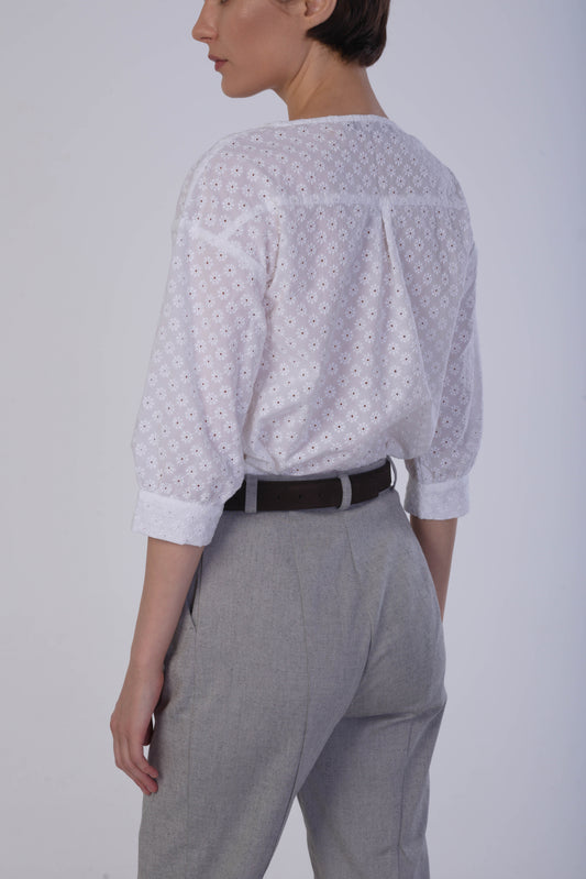 A woman shown from the back wearing a white eyelet embroidered blouse with three-quarter sleeves and a dark brown belt, paired with light gray trousers, against a plain light gray studio background.