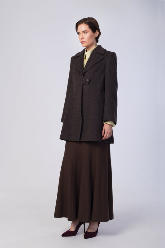 A model stands in a studio wearing a brown tailored coat over a pale yellow top, paired with a matching dark brown maxi skirt and dark burgundy heels, against a light gray background.