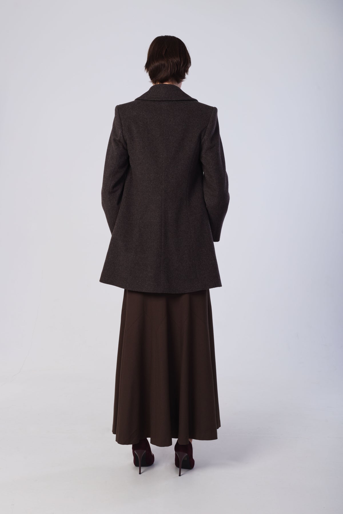 A model viewed from the back wearing a dark brown textured coat and a matching dark brown maxi skirt, paired with dark pointed-toe heels, against a light gray studio background.