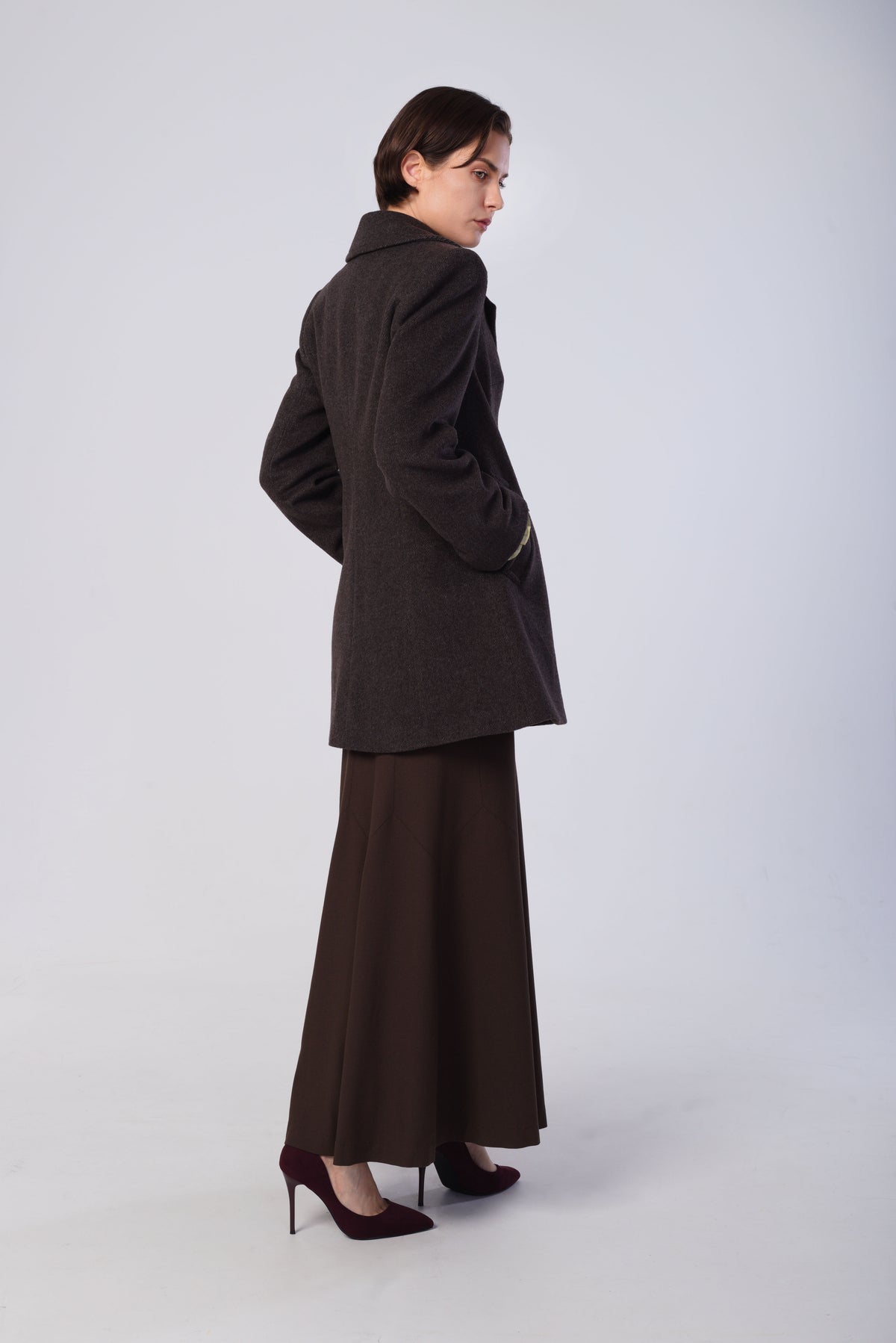A model is shown from a side-back angle wearing a dark brown wool-blend coat, a matching dark brown wide-leg skirt, and dark burgundy high heels against a light gray studio background.