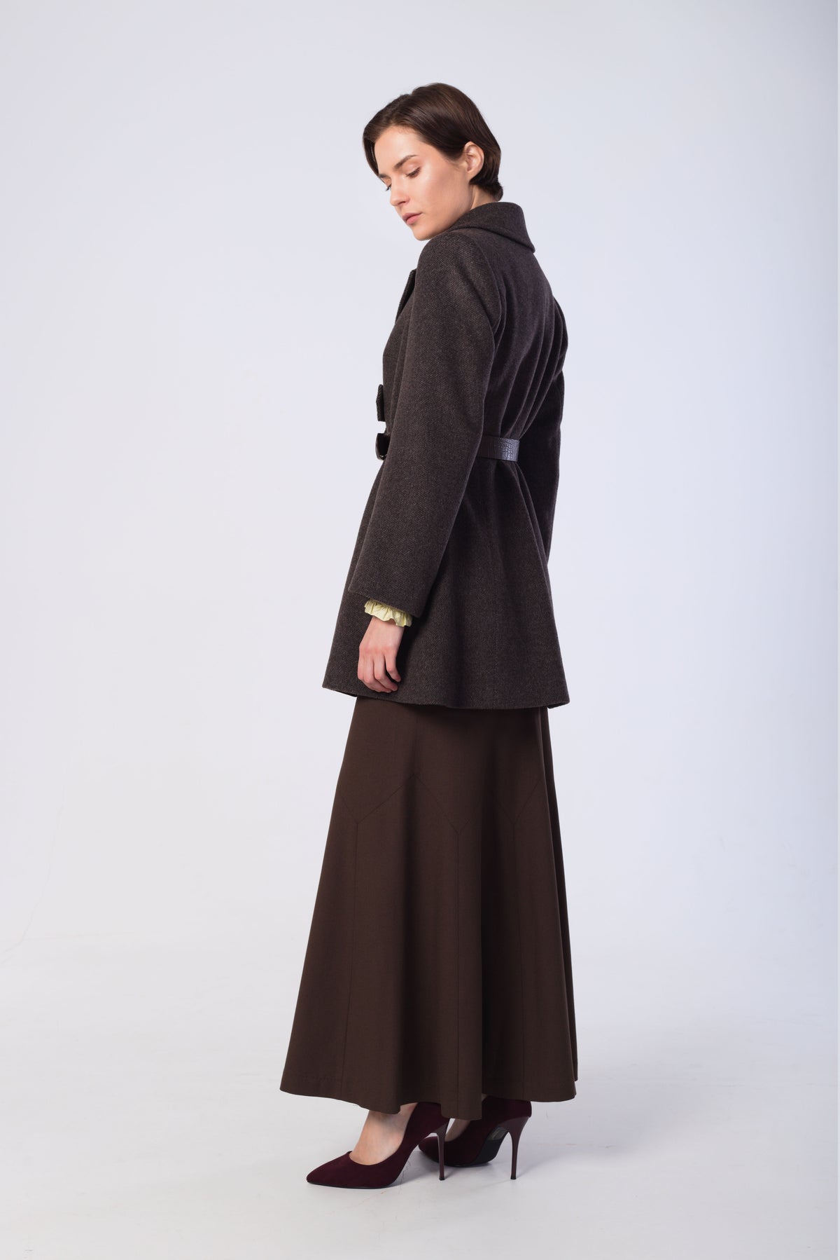 A woman modeling a dark brown coat with a matching long skirt and dark heels, photographed against a light gray studio background.