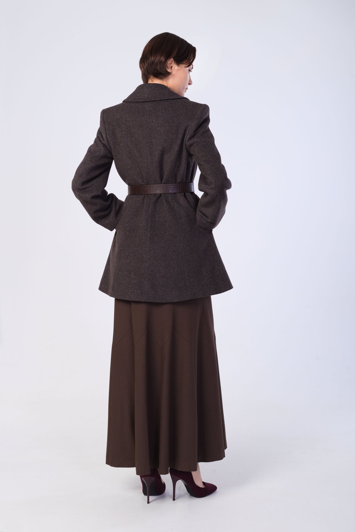 A woman shown from the back wearing a dark brown tailored coat with a matching belt and a long brown skirt, photographed against a plain light gray studio background.