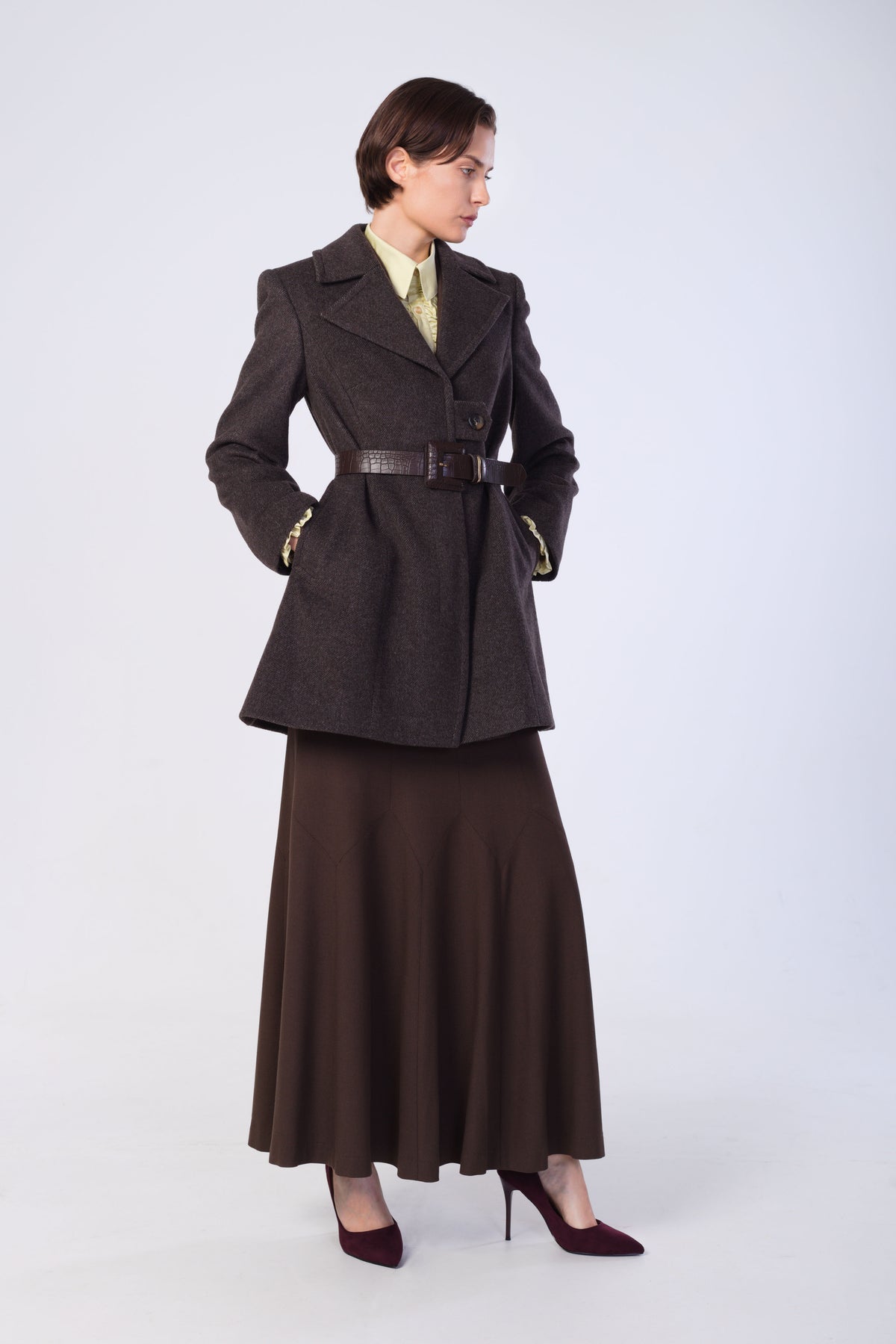 A model wearing a dark brown belted coat over a matching brown skirt, paired with a light green collared shirt and burgundy heels, photographed against a light gray studio background.