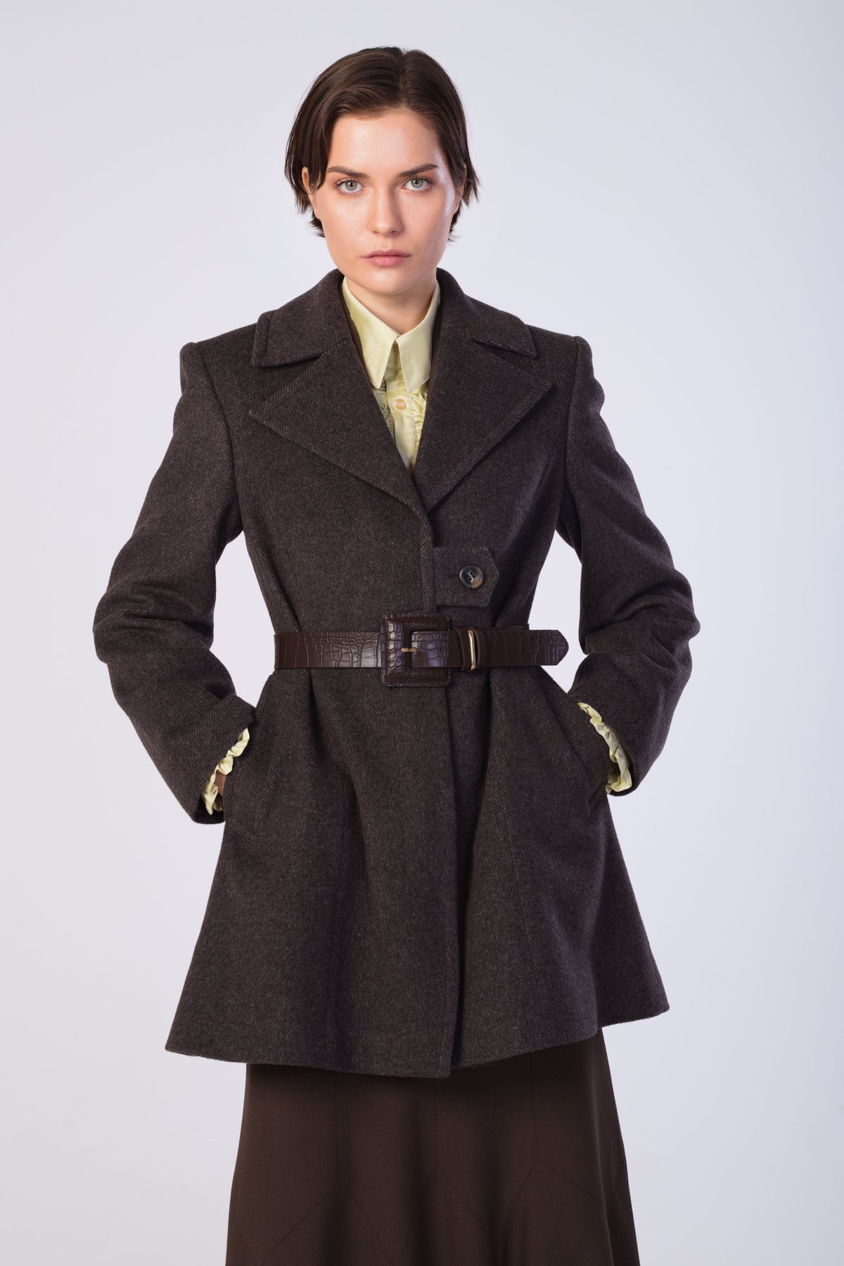 A model wearing a brown wool-blend coat with a matching belt, styled over a pale yellow shirt and dark skirt, against a light gray studio background.