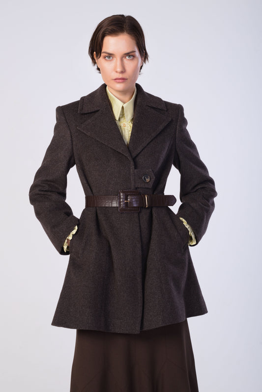 A model wearing a brown wool-blend coat with a matching belt, styled over a pale yellow shirt and dark skirt, against a light gray studio background.