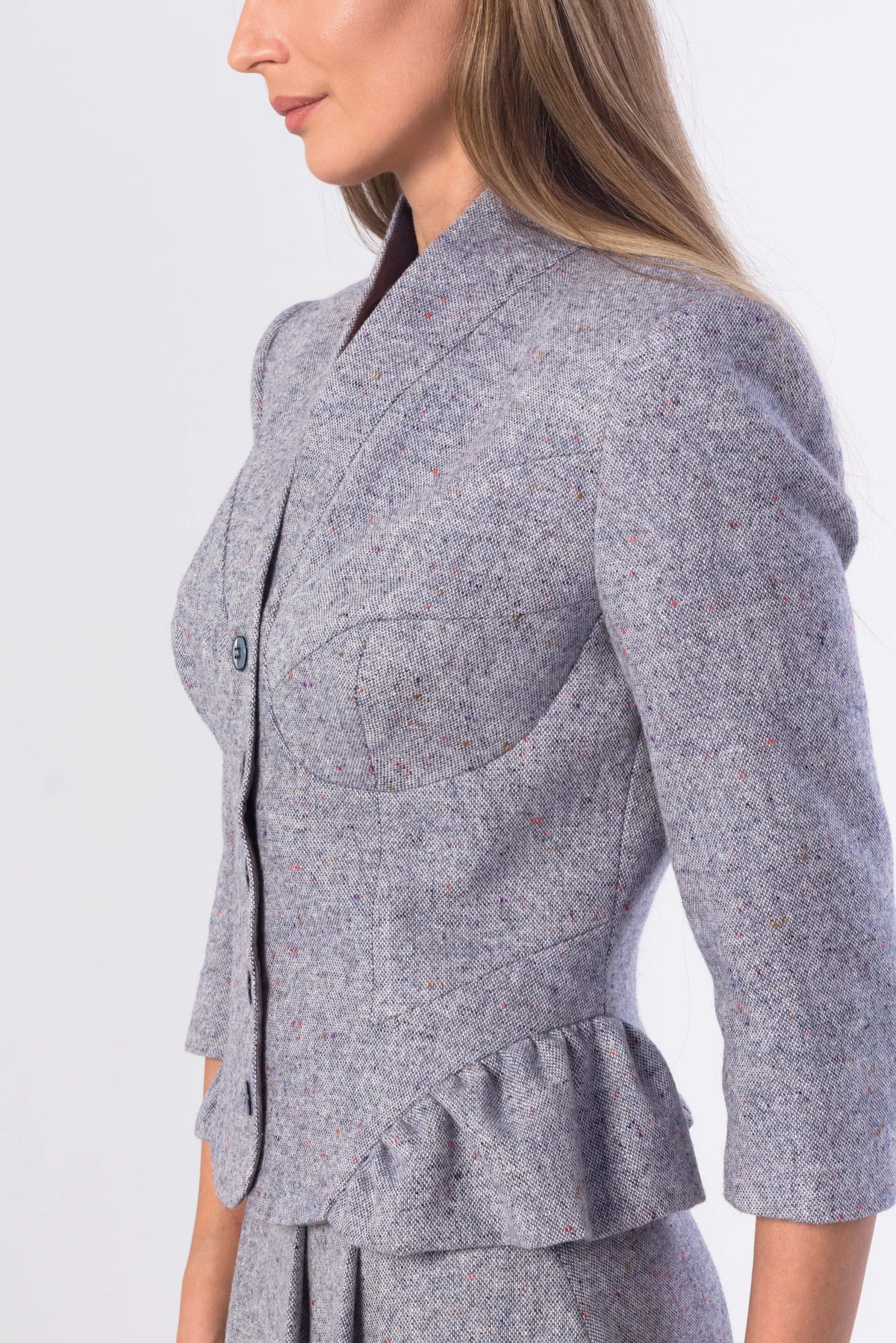 A woman wearing a structured light blue-grey tweed blazer with subtle multicolor flecks, shown in a side profile against a light gray studio background.