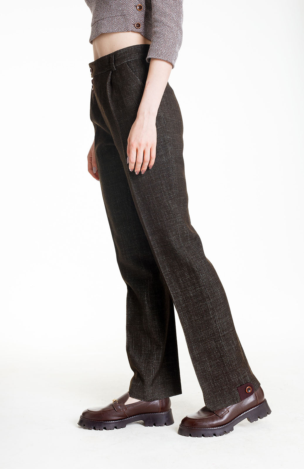 A side angle shot focusing on the lower half of the trousers. The deep olive fabric is paired with a cropped grey herringbone top. The trousers feature a sharp front crease and a distinctive maroon button-tab detail at the outer side seam of the hem.