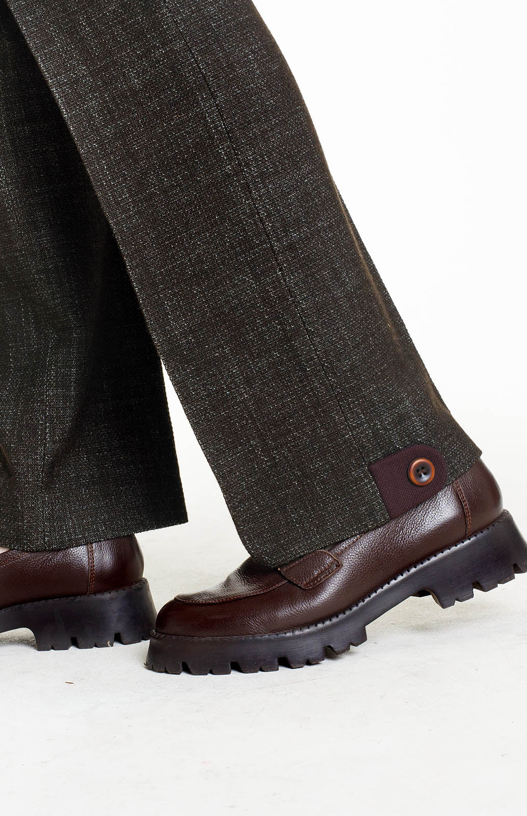 A close-up detail shot of the trouser hem resting on dark brown platform loafers. It highlights the maroon contrast fabric "half-belt" tab with a single button at the side seam, adding an architectural touch to the straight-leg finish.