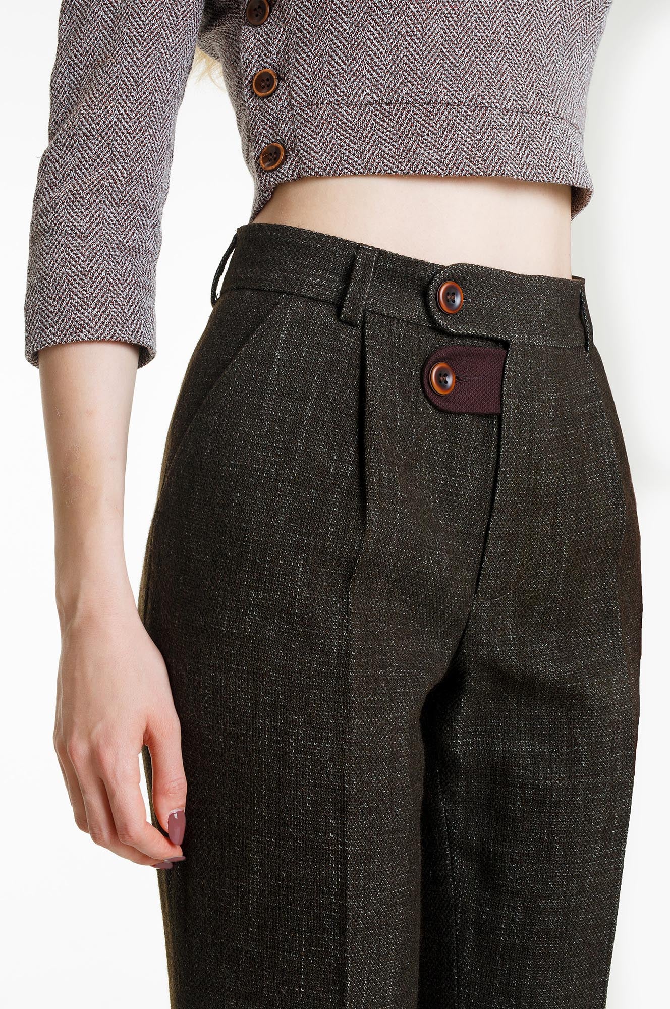 A detailed close-up of the high waistband. The shot focuses on the textured olive material and the maroon contrast tab at the fly, secured with two brown buttons. The trousers are paired with a grey cropped top, emphasizing the high-rise tailoring.