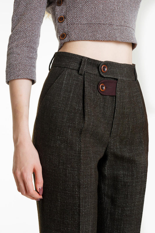 A detailed close-up of the high waistband. The shot focuses on the textured olive material and the maroon contrast tab at the fly, secured with two brown buttons. The trousers are paired with a grey cropped top, emphasizing the high-rise tailoring.
