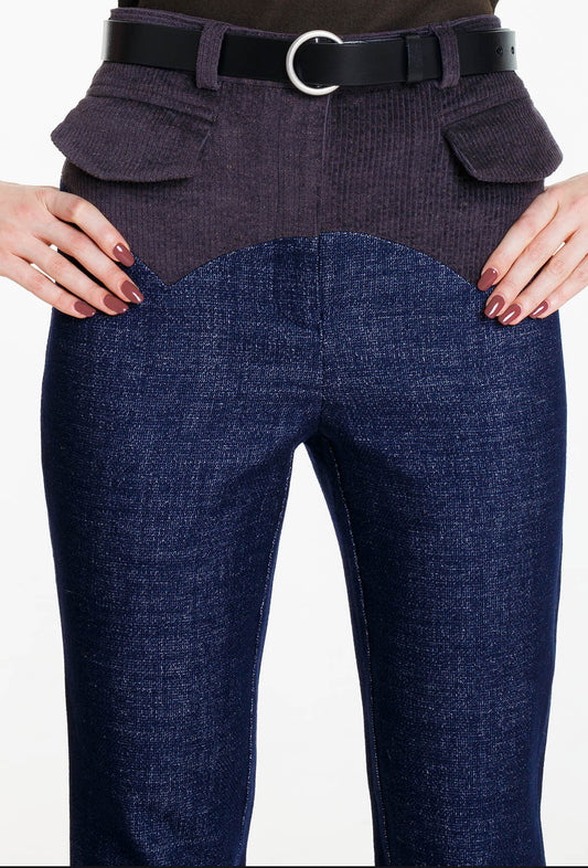 A symmetrical close-up of the waistband and yoke area. The pointed anthracite corduroy panel and the two front flap pockets against the deep blue wool denim. The woman's hands with red nails rest on the hips.
