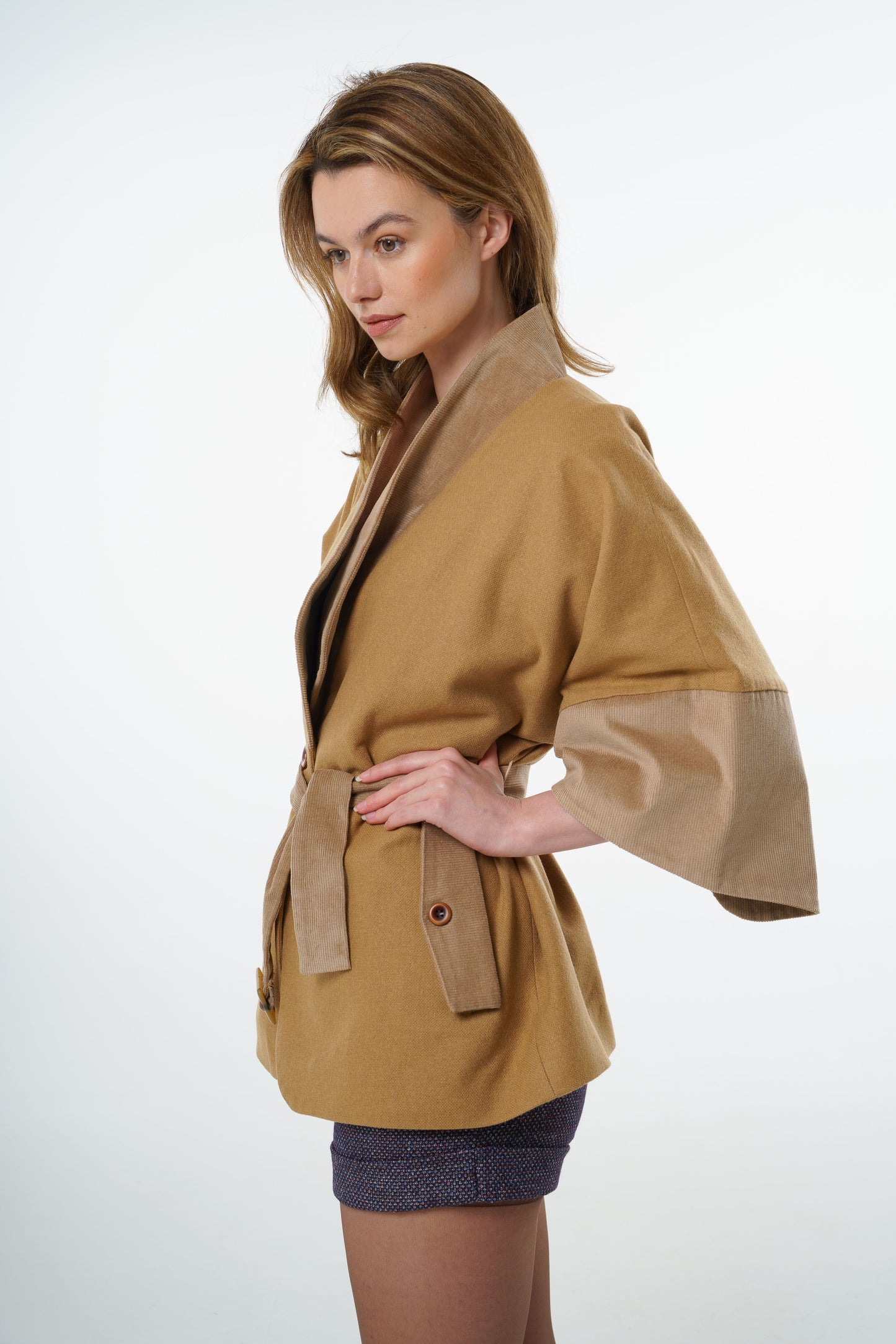 Profile shot of a tan-beige belted jacket, worn by a model, emphasizing the structured wrap silhouette and earthy tones.