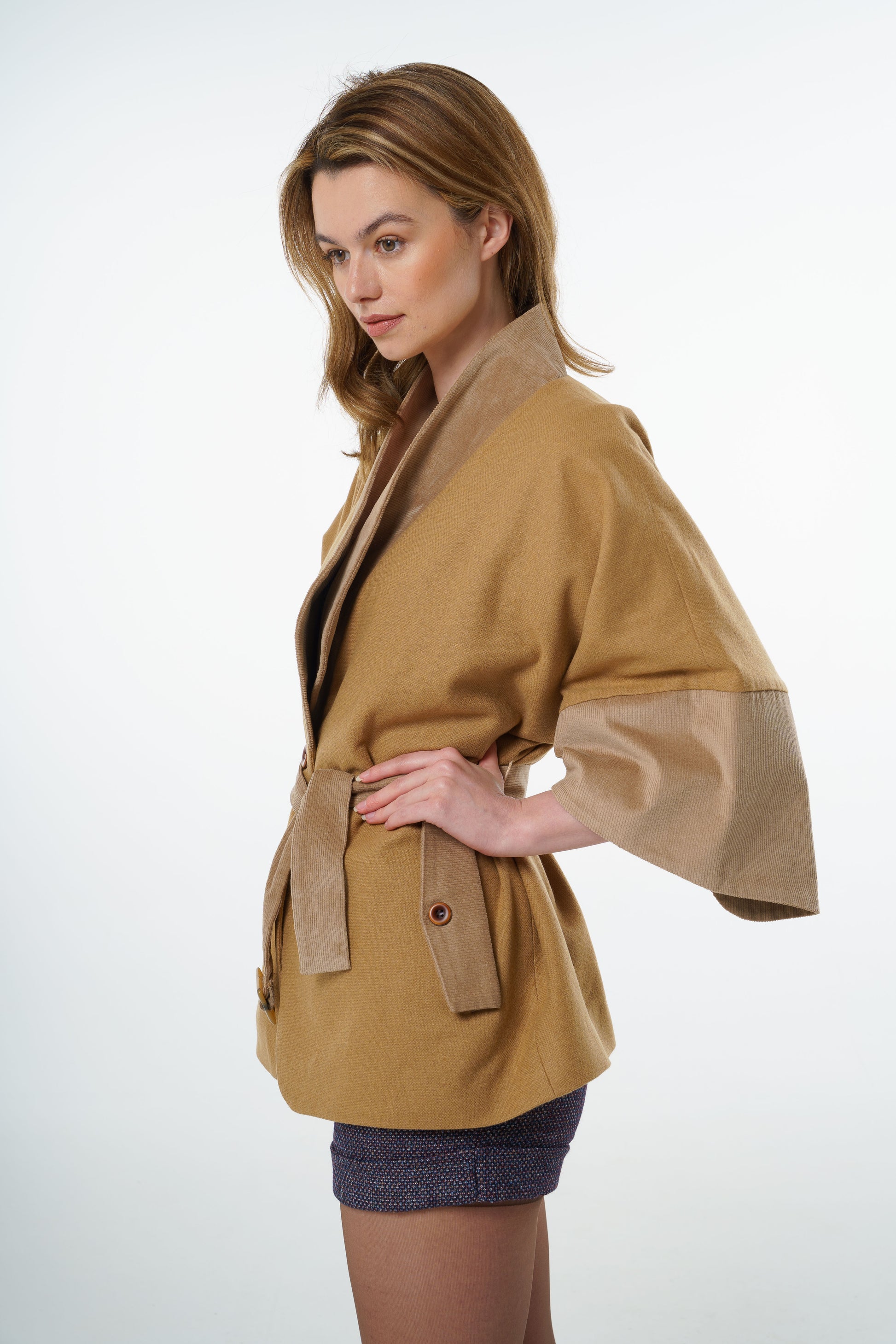 Profile shot of a tan-beige belted jacket, worn by a model, emphasizing the structured wrap silhouette and earthy tones.