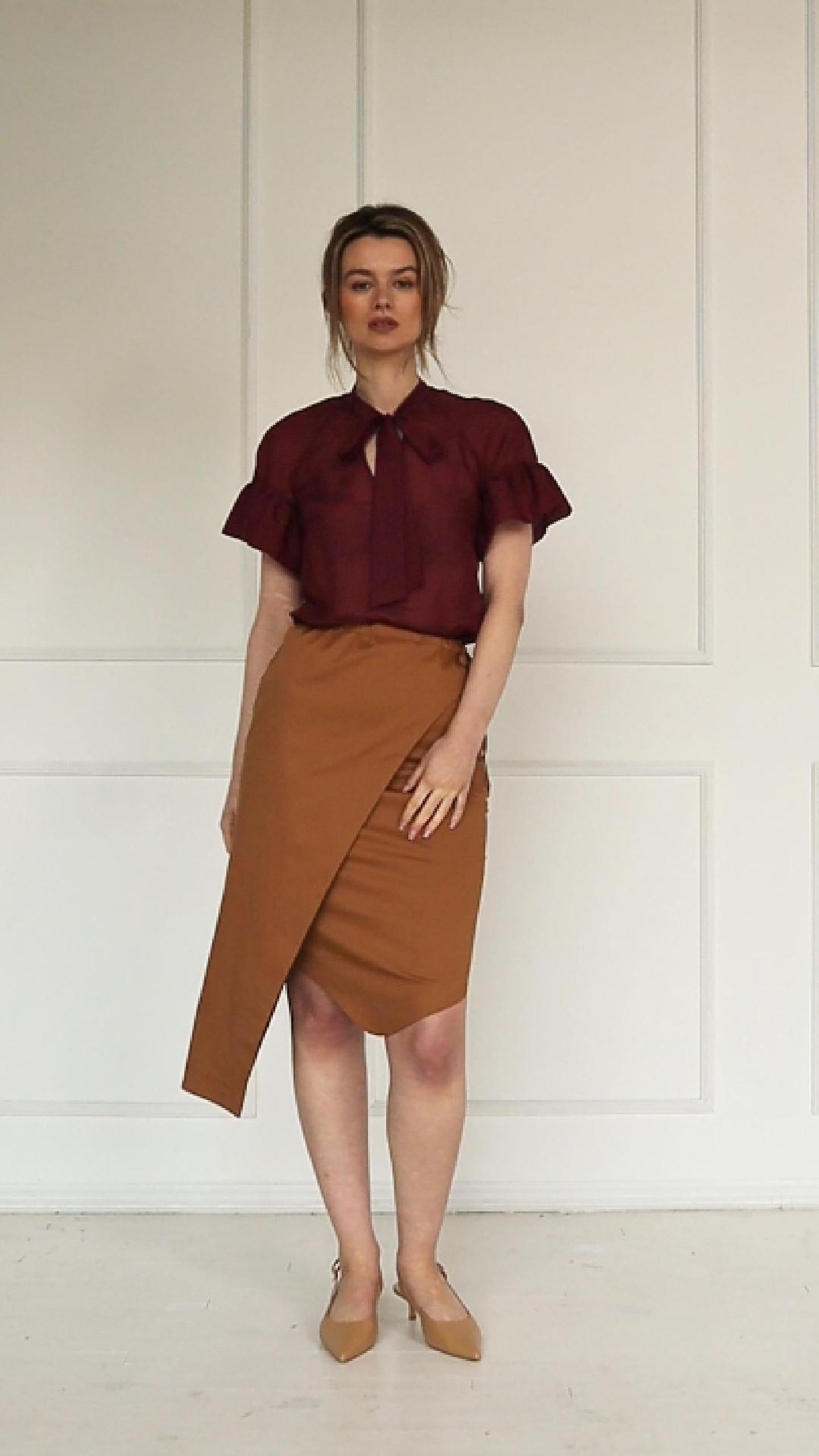 A studio video showing a camel wrap pencil skirt, highlighting the asymmetric overlay, side tie, and a burgundy cotton–silk raglan‑sleeve blouse, wearing by a model, highlighting the neck tie, ruffled cuffs, and fluid drape of the fabric.